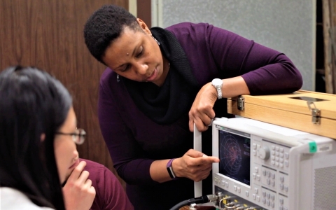 A photo of Dr. Rhonda Franklin, dressed in a purple long sleeve shirt and black scarf, pointing at the screen of an instrument. She is pointing out something of interest to a nearby seated student.
